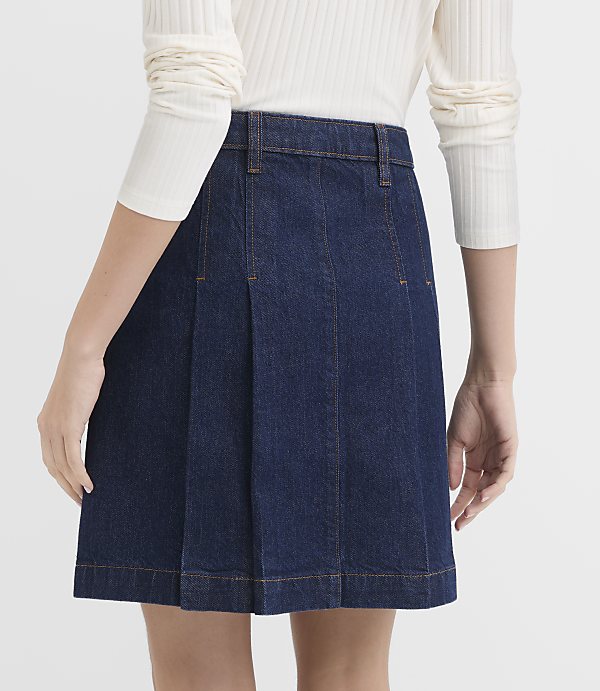 A person wearing a dark denim pleated skirt and a white long-sleeved shirt stands against a plain white background.