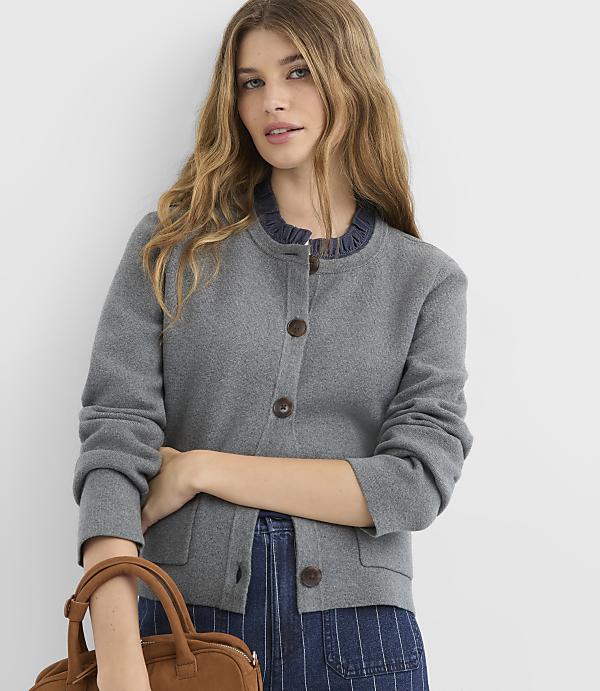 A young woman with long, wavy blonde hair stands in the foreground wearing a grey cardigan and blue striped pants, with a brown handbag at her side, against a plain white background.
