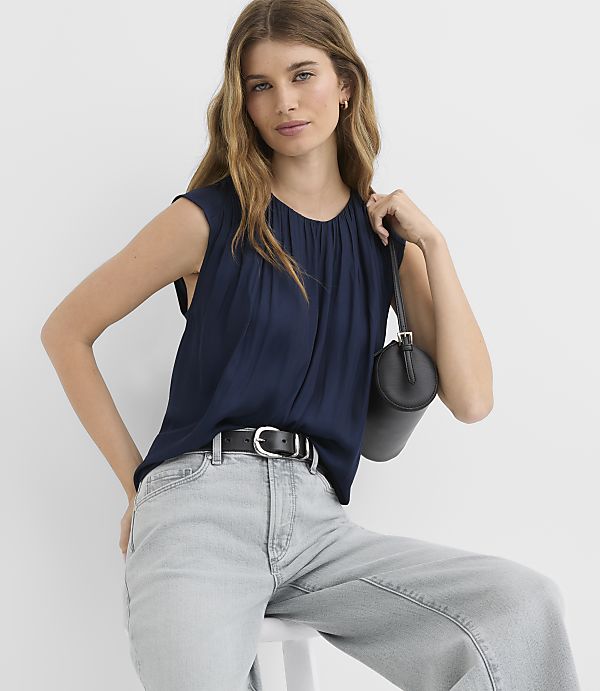 A woman in a navy blue top and light gray jeans poses with a black handbag against a plain white background.