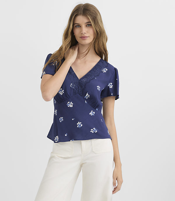 A young woman in a floral navy blue blouse and white pants stands against a plain white background.