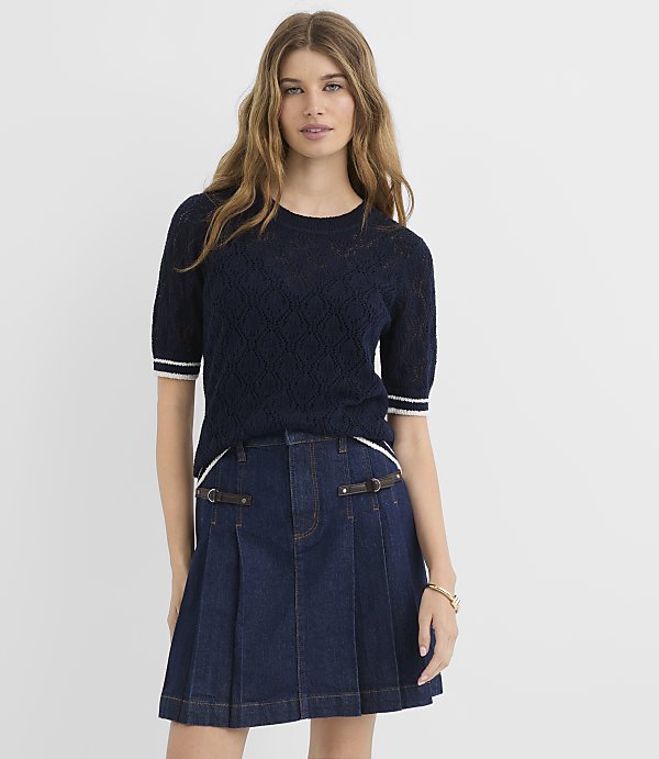 A woman in a navy blue textured sweater and a dark denim pleated skirt stands against a plain white background.