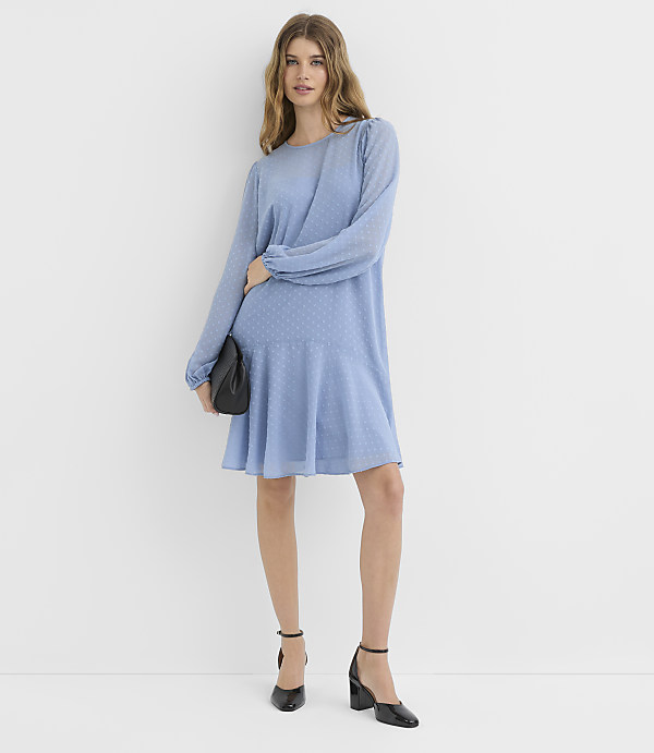 A woman in a light blue dress and black heels stands against a plain white background, holding a black clutch.