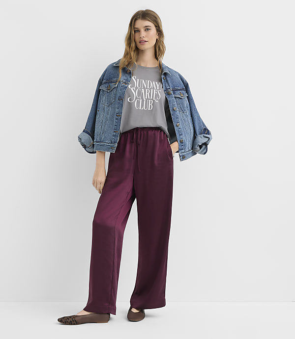 A woman in a denim jacket and burgundy satin pants stands against a plain white background.