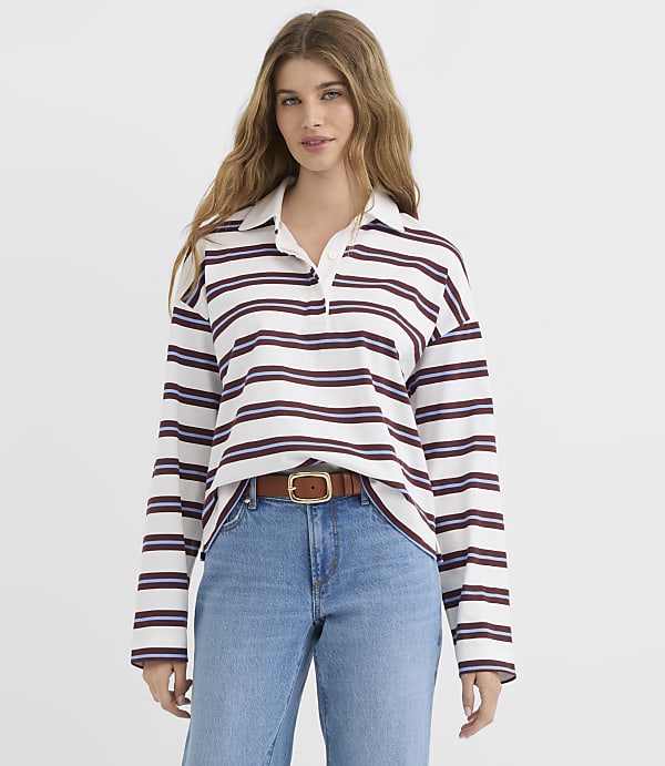 A young woman with long, wavy brown hair stands in the foreground wearing a striped long-sleeved shirt and blue jeans, with a plain white background behind her.