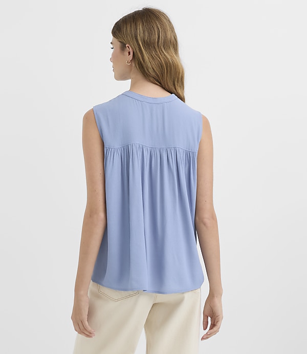 A woman in a blue sleeveless top and cream pants stands against a plain white background.