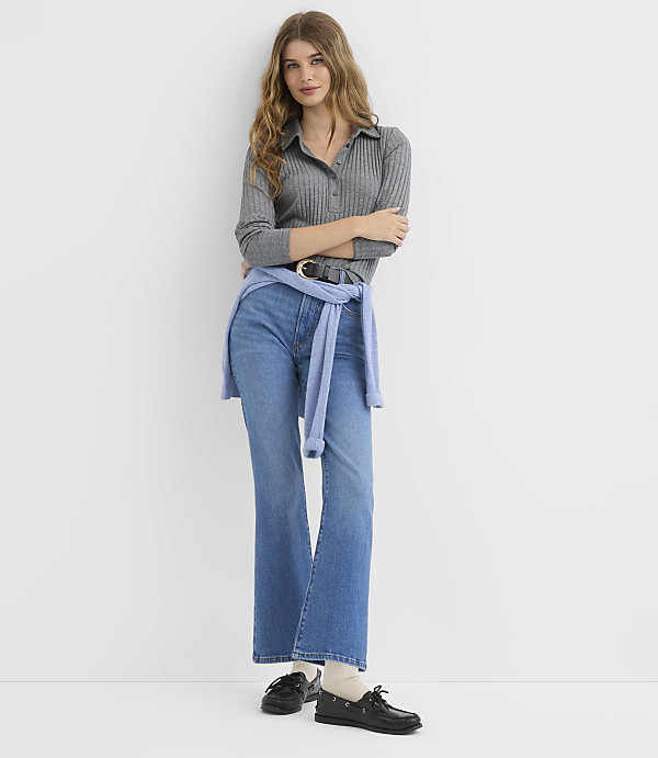 A woman in a grey ribbed polo shirt and blue jeans stands against a plain white background, with a light blue sweater tied around her waist.