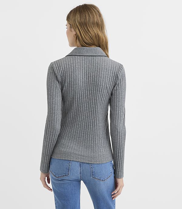 A woman in a grey ribbed sweater and blue jeans stands against a plain white background.