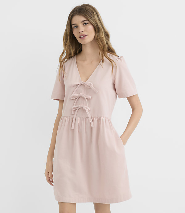 A woman in a pink dress with bow details stands against a plain white background.