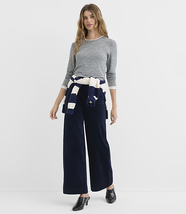 A woman in a gray long-sleeved shirt and navy wide-leg pants stands against a plain white background, with a striped sweater tied around her waist.