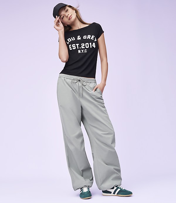 A woman in a black t-shirt and grey pants poses against a plain purple background.