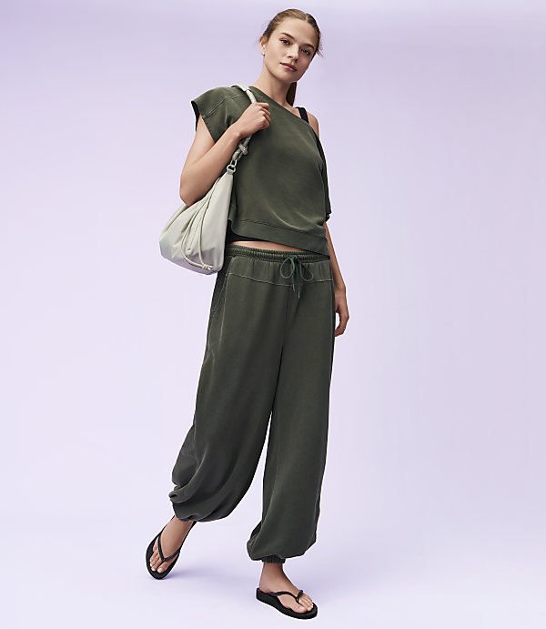 A woman in a green outfit and flip-flops stands with a bag over her shoulder against a light purple background.