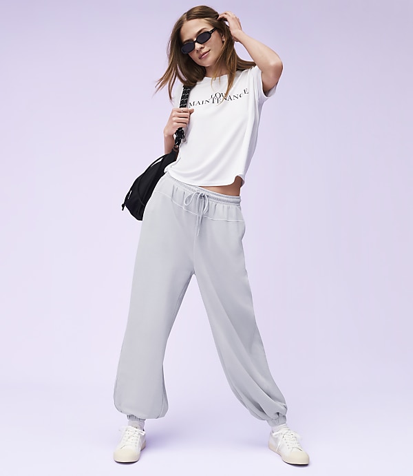 A woman in a white t-shirt and light blue sweatpants poses against a soft purple background.