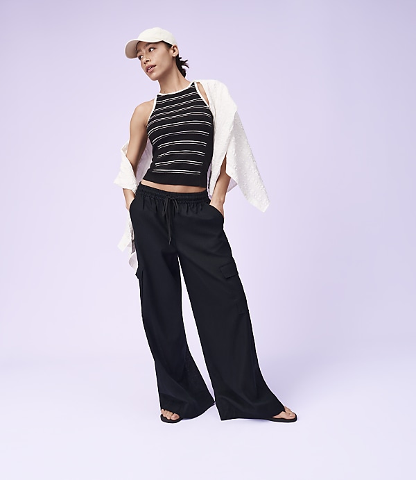 A woman in a striped tank top, black cargo pants, and a white cap stands against a plain purple background.