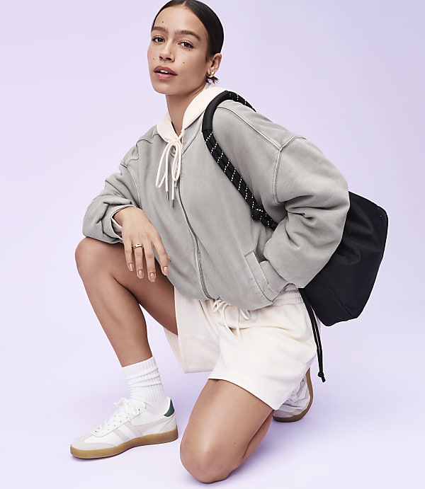 A woman in athletic wear crouches in the foreground with a black bag slung over her shoulder, against a plain purple background.