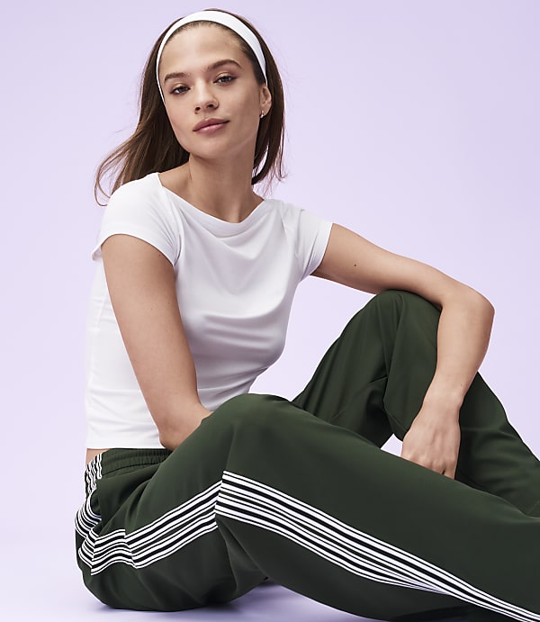 A young woman in a white t-shirt and green track pants with white stripes sits against a soft purple background.