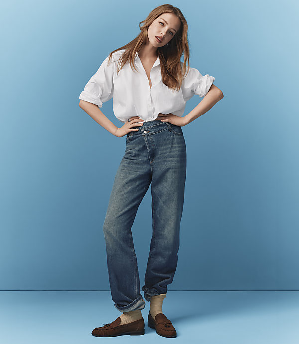 A young woman in a white shirt and blue jeans stands with her hands on her hips against a solid blue background.