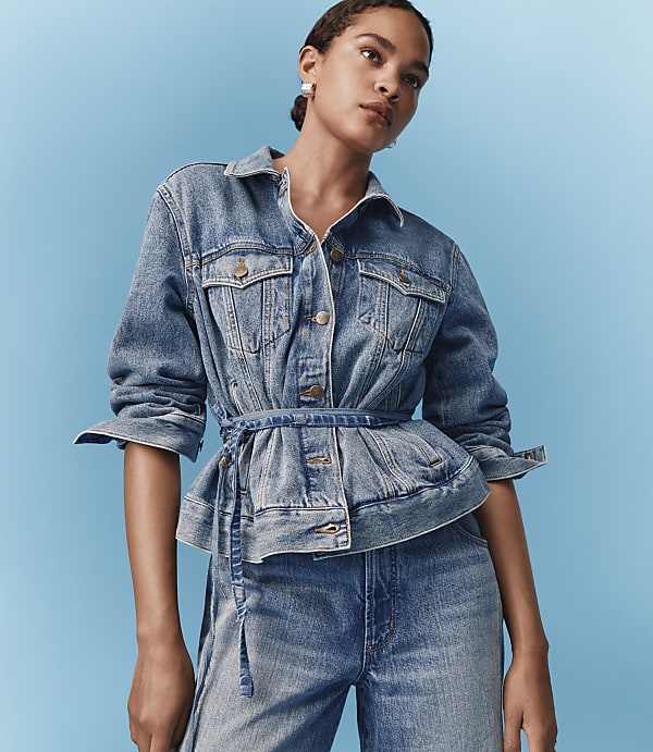 A woman in a denim jacket and jeans stands against a bright blue background.