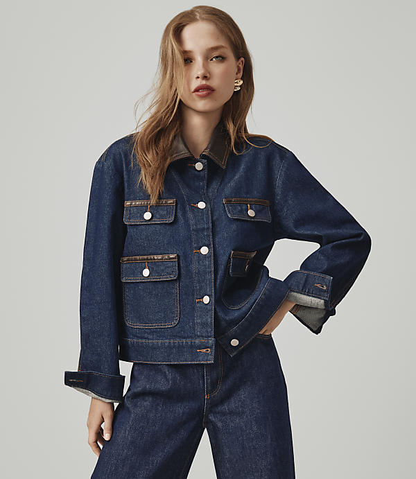 A young woman in a denim jacket and jeans stands against a plain gray background.
