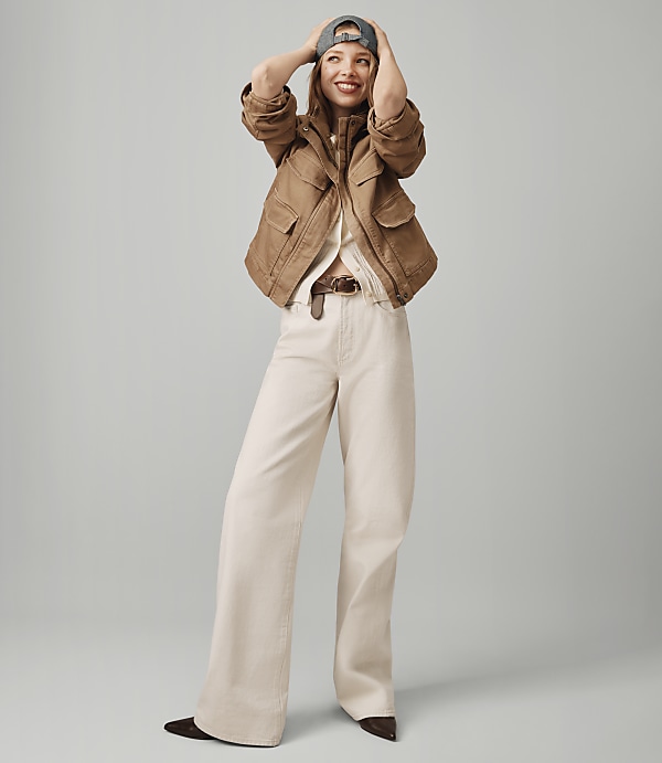 A smiling woman in a brown jacket and cream pants stands against a plain gray background.