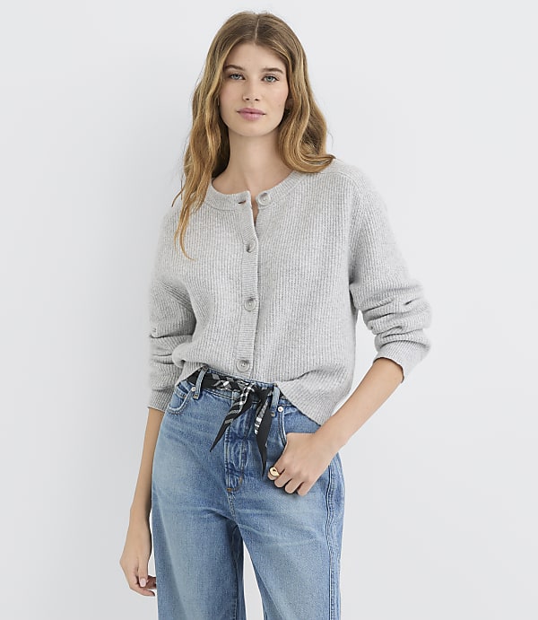 A woman in a grey cardigan and blue jeans stands against a plain white background.