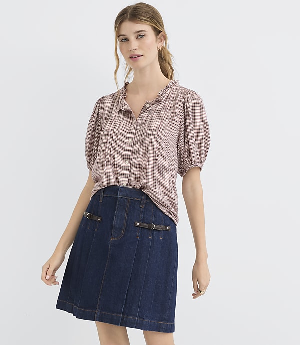 A young woman in a ruffled, checkered blouse and a dark denim skirt stands against a plain white background.