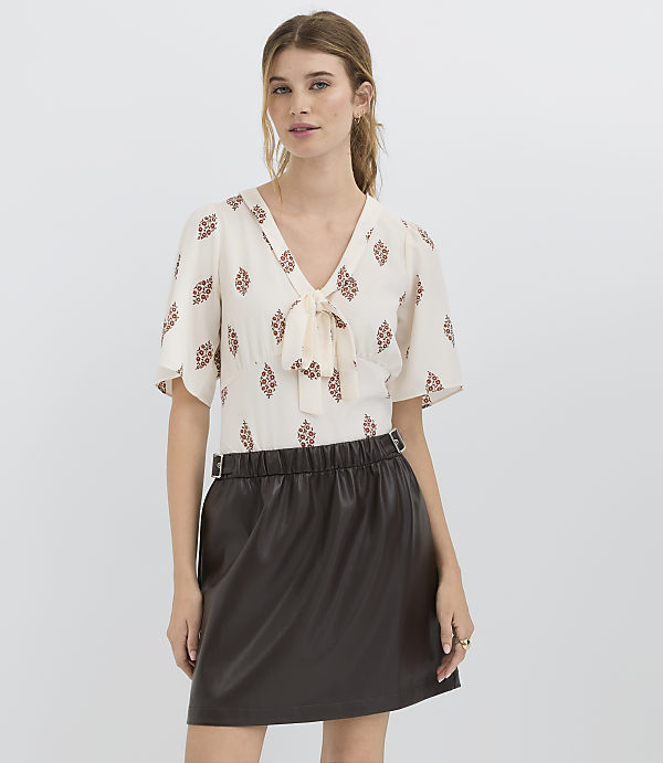 A woman in a patterned blouse and a dark skirt stands against a plain white background.