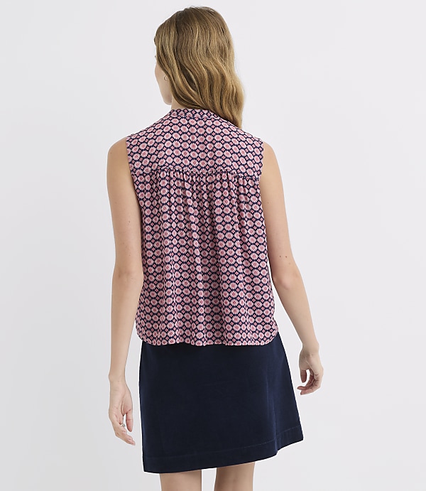 A woman in a patterned sleeveless top and a dark skirt stands against a plain white background.