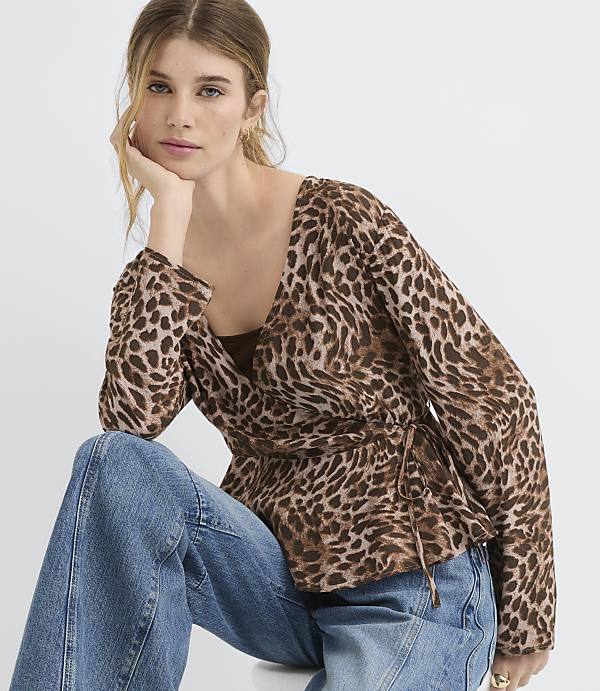 A woman in a leopard-print wrap top and blue jeans poses against a plain white background.