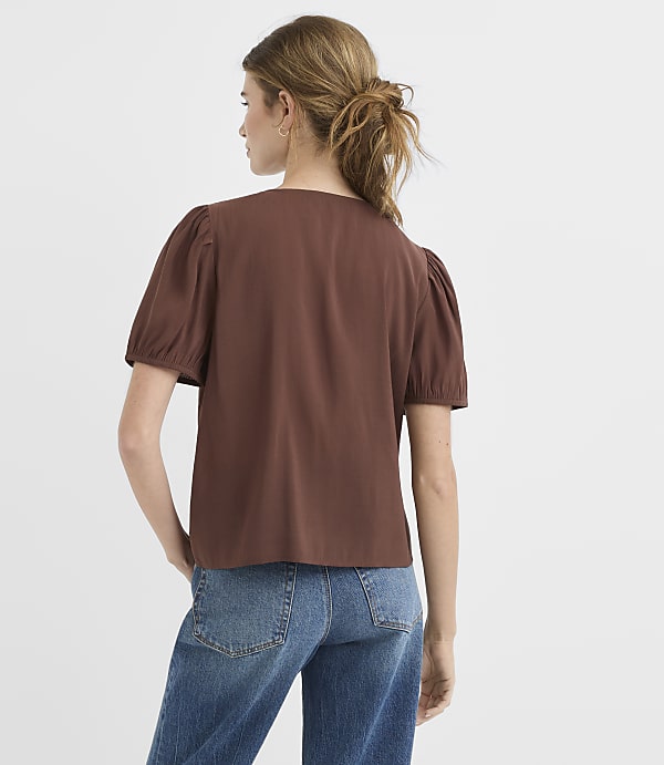 A woman in a brown puff-sleeve top and blue jeans stands against a plain white background.