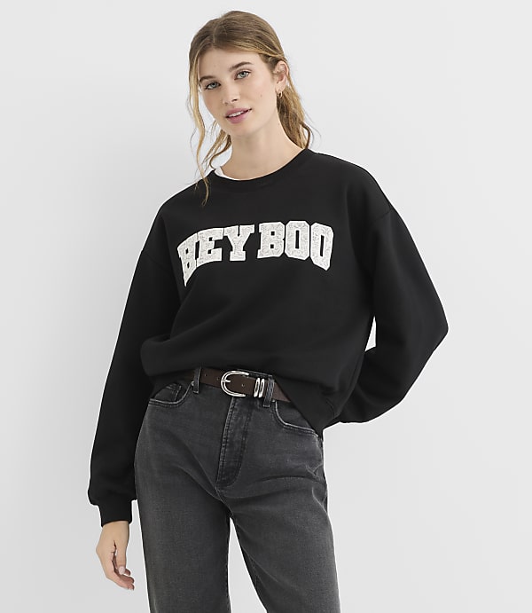 A young woman in a black sweatshirt with "HEY BOO" printed on it and dark jeans stands against a plain white background.