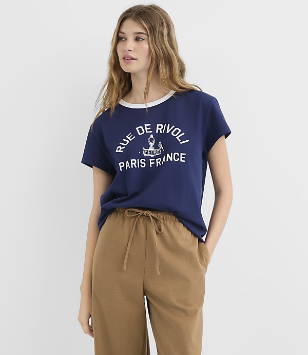 A young woman in a navy t-shirt with "Rue de Rivoli Paris France" printed on it and brown drawstring pants stands against a plain white background.