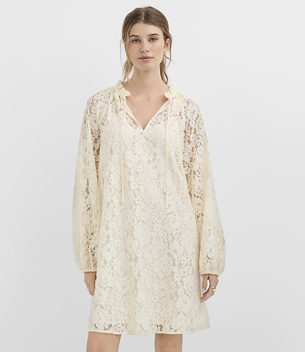 A woman in a cream lace dress stands against a plain white background.