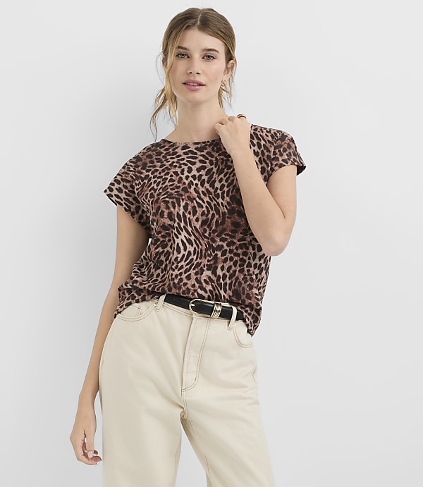 A woman in a leopard print t-shirt and cream pants stands against a plain white background.
