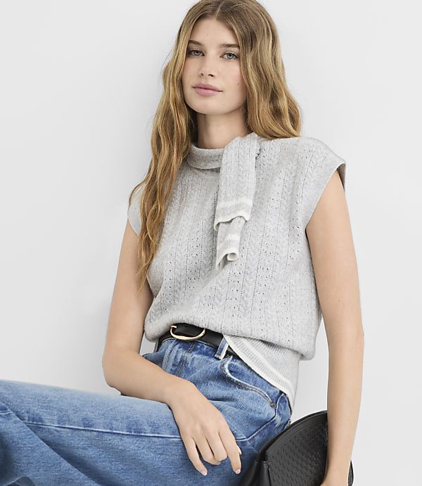 A young woman with long blonde hair, wearing a grey sleeveless turtleneck sweater and blue jeans, sits against a plain white background.