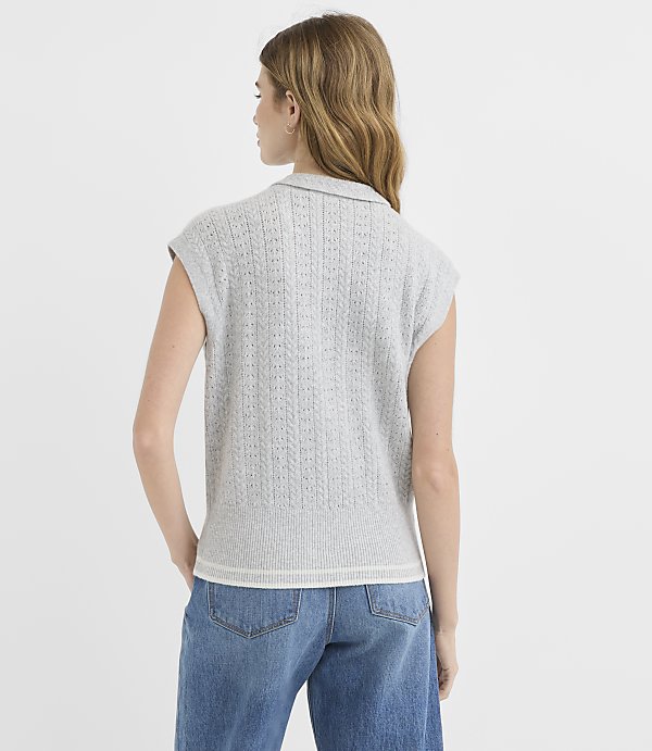 A woman in a gray sleeveless sweater and blue jeans stands against a plain white background.