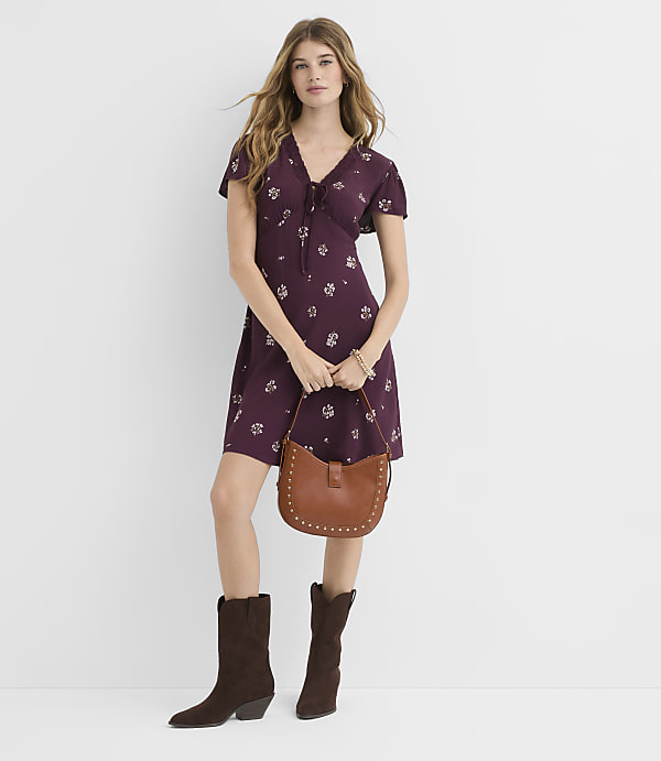 A woman in a floral dress and cowboy boots stands against a plain white background, holding a brown studded purse.