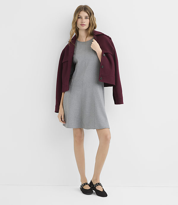 A woman in a gray dress and maroon jacket stands against a plain white background.