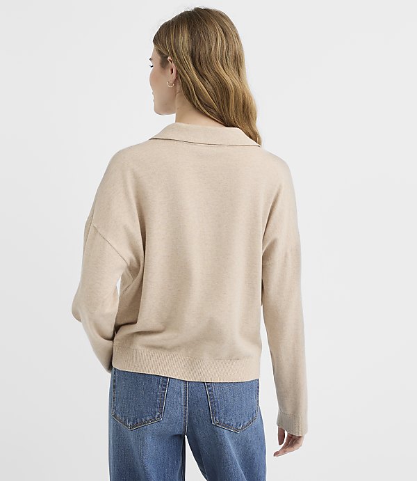 A woman in a beige collared sweater and blue jeans stands against a plain white background.