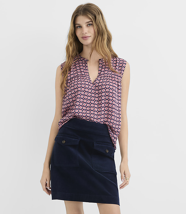 A woman in a patterned top and navy skirt stands against a plain white background.