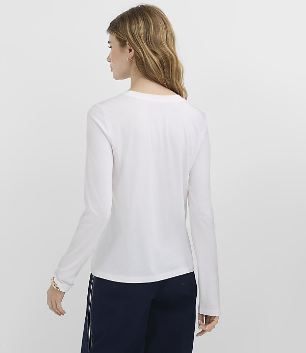 A woman in a white long-sleeved shirt and dark pants stands against a plain white background.