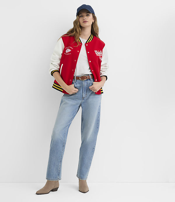 A woman in a red and white varsity jacket and light-wash jeans stands against a plain white background.