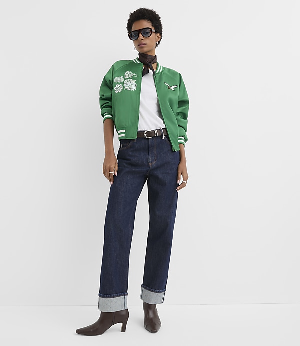 A woman in a green bomber jacket and dark jeans stands against a plain white background.