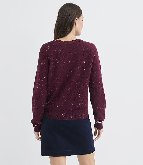A woman in a burgundy speckled sweater and navy skirt stands against a plain white background.