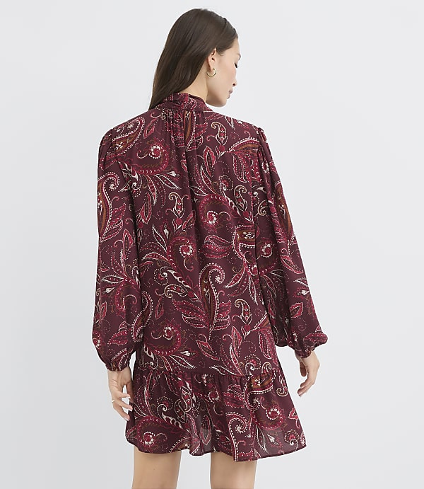 A woman in a burgundy paisley dress with a ruffled hem stands against a plain white background.