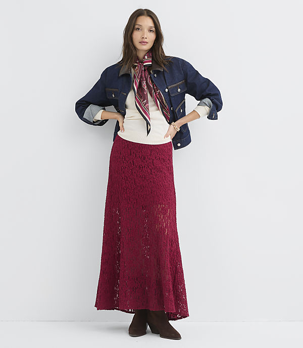 A woman in a denim jacket and a long red skirt stands against a plain white background.