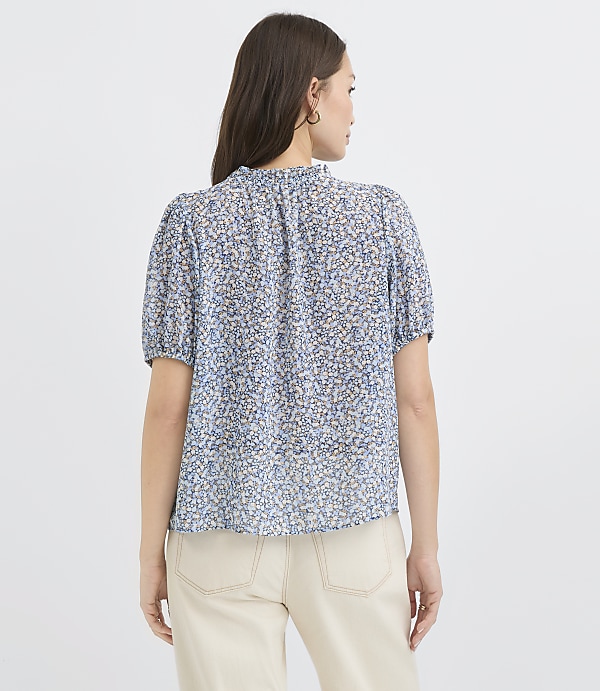 A woman in a blue floral blouse and cream pants stands against a plain white background.