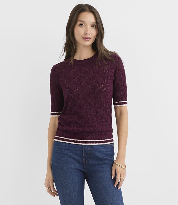 A woman in a burgundy, short-sleeved, textured sweater with striped cuffs and hem stands against a plain white background.