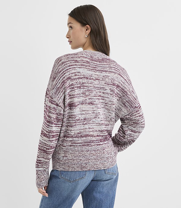 A woman in a marled burgundy and grey sweater and blue jeans stands against a plain white background.
