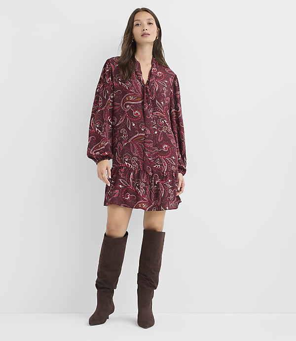 A woman in a paisley print dress and brown boots stands against a plain white background.