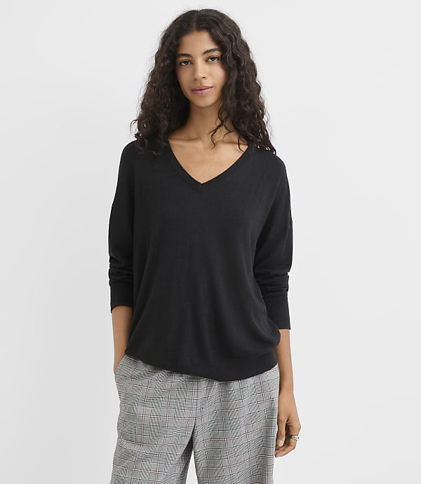 A woman in a black v-neck sweater and plaid pants stands against a plain white background.